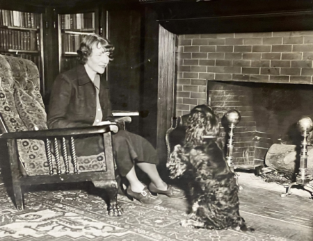 Esther Forbes seated in front of a fireplace with a dog facing her.